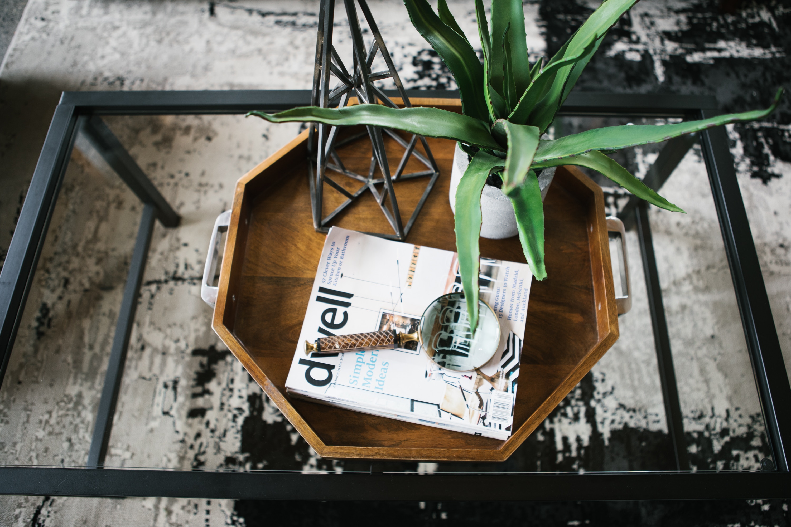 overhead view of a glass coffee table with a small aloe vera plant in a white pot beside an architecture design magazine and a magnifying glass