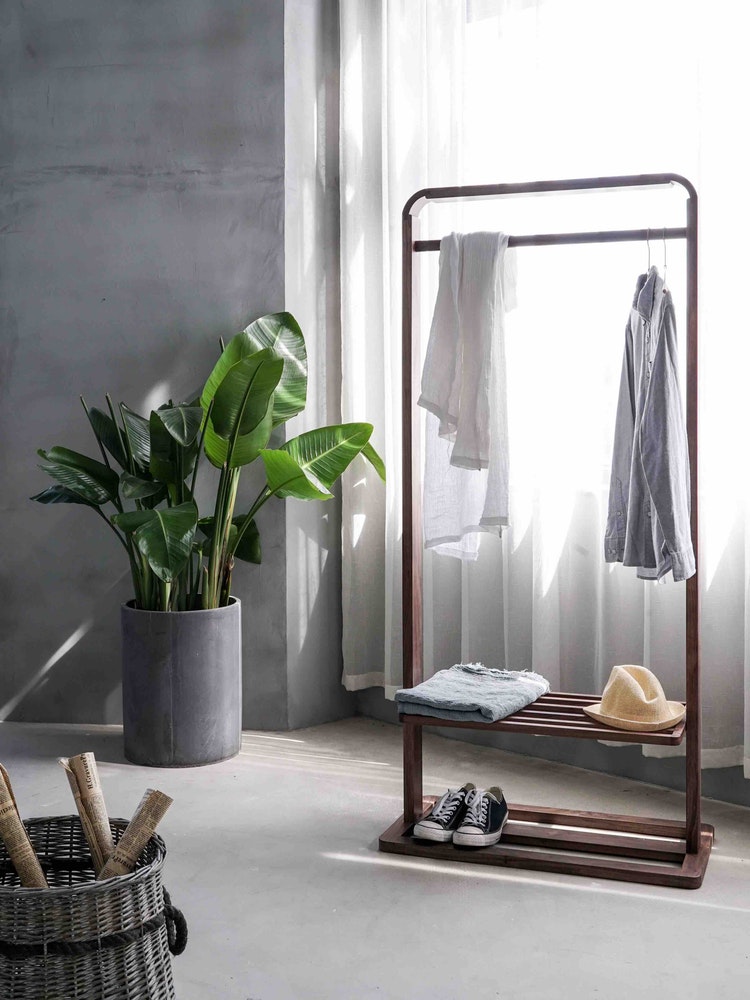 a large birds of paradise plant in a silver planter beside a wooden clothing rack in a modern concrete room